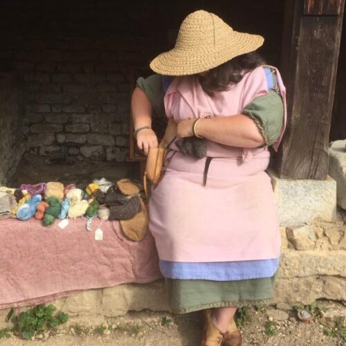 Tutor Sue Day at Chedworth Villa, wearing a green and blue dress, pink apron and wide brimmed straw hat with her naturally dyed yarn beside her